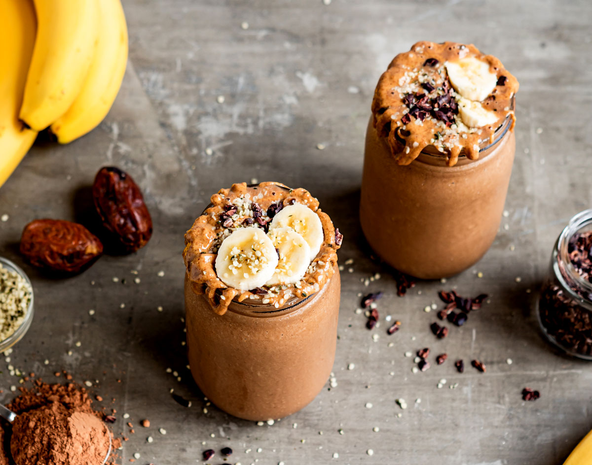 Two glasses of chocolate protein shake with sliced bananas and cacao nibs on tip.