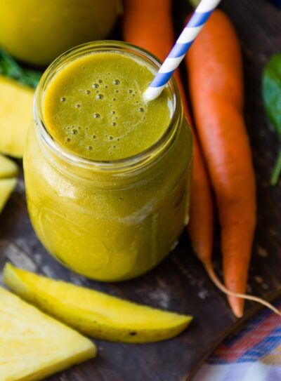 Carrot greens smoothie in a mason jar with a paper straw next to carrot tops on table.