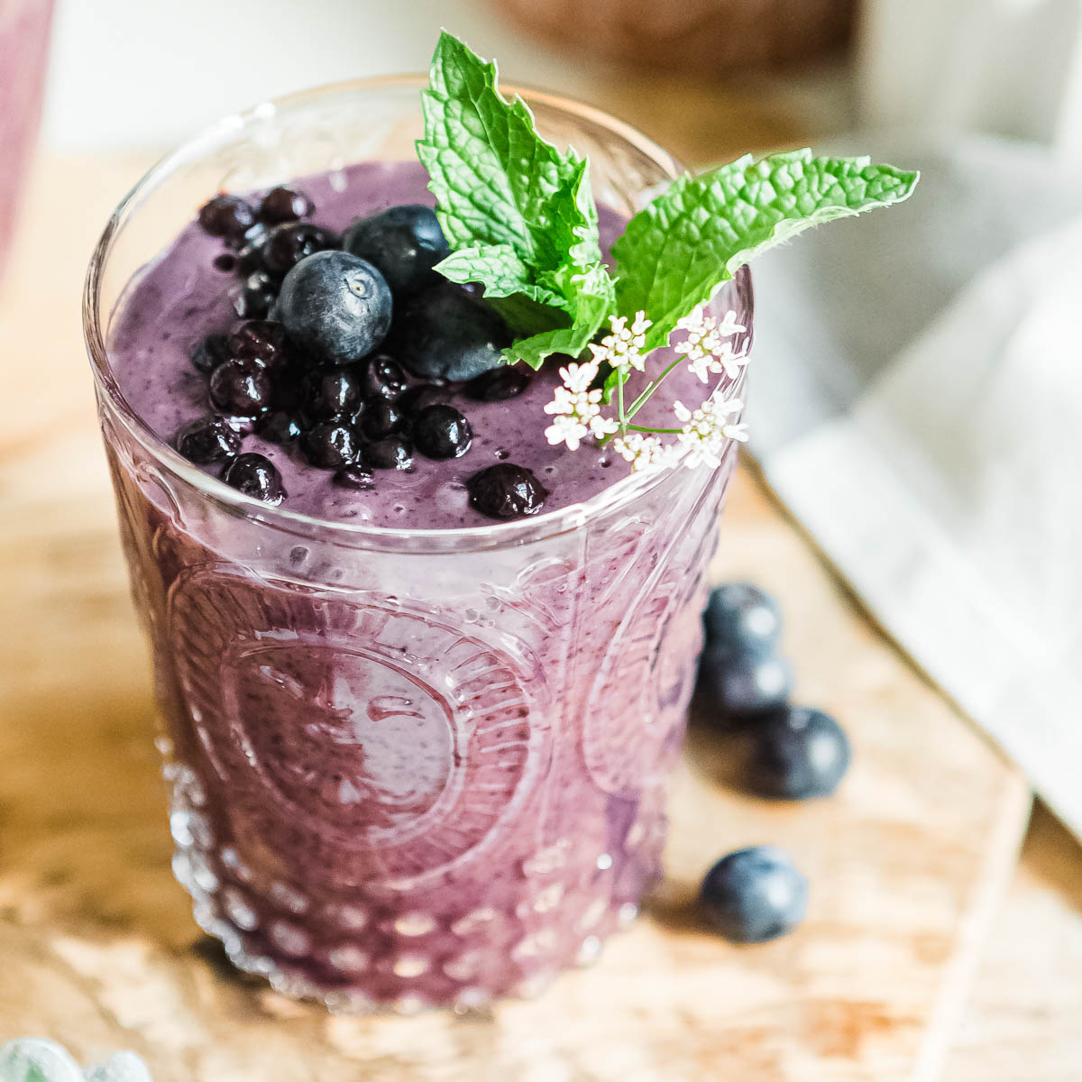 Glass of blueberry smoothie on a woodcutting board surrounded by fresh blueberries.