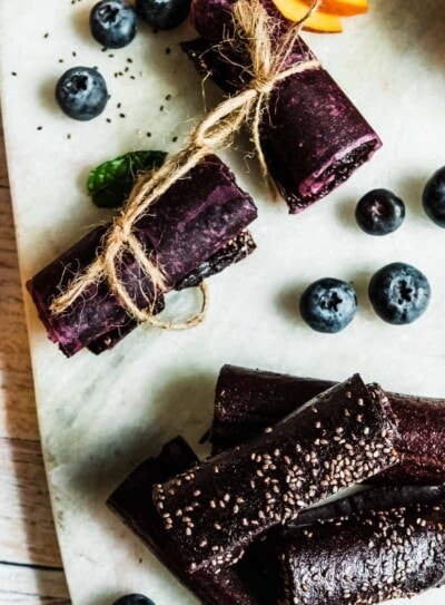rolls of fruit leather on a marble serving tray surrounded by fresh blueberries and peaches.