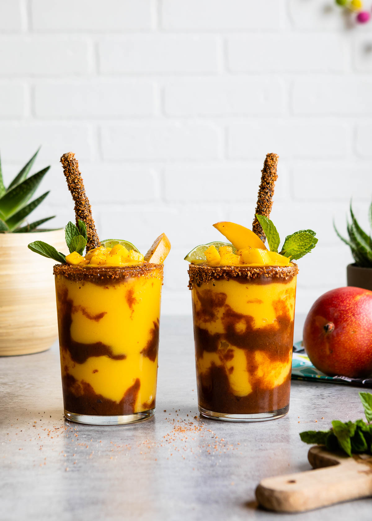 Two glasses filled with Mexican mangonada recipe garnished with fresh fruit and a Tahin dipped straw.