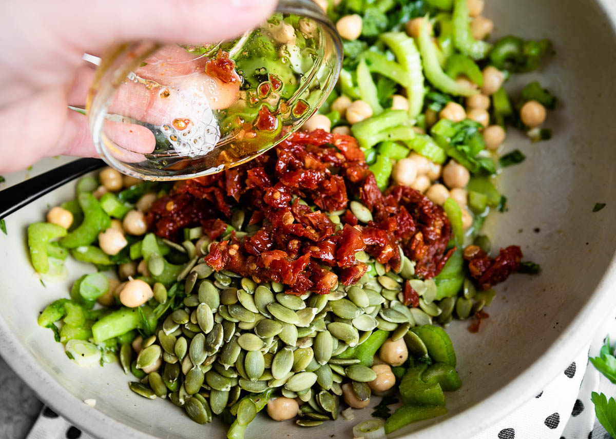 pouring sun-dried tomatoes and pepitas into a white bowl with other green ingredients.