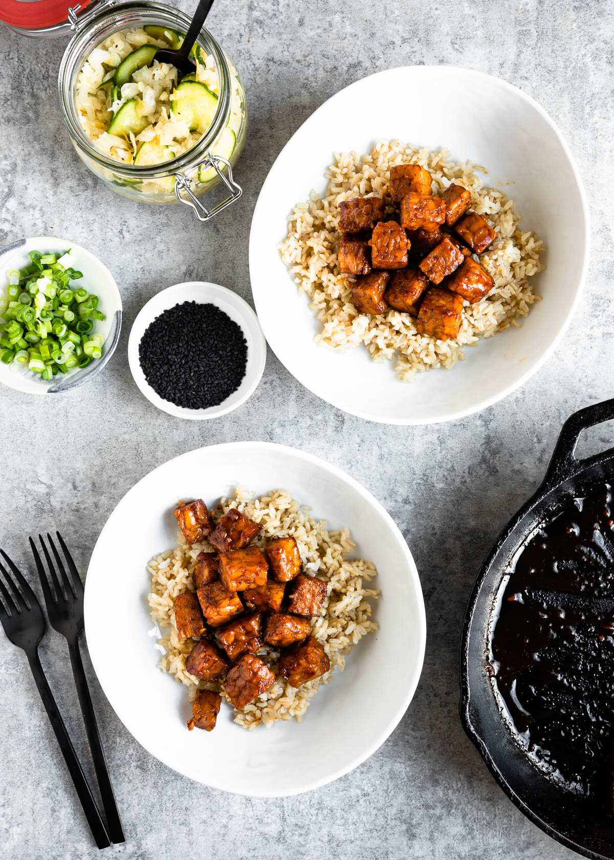 Bowls with brown rice and cooked tempeh next to small bowls of green onion, sesame seeds and pickled vegetables.