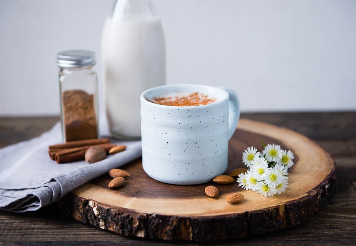 A mug of warm almond milk placed on a beautiful wooden cutting board, surrounded by natural ingredients and flowers. 